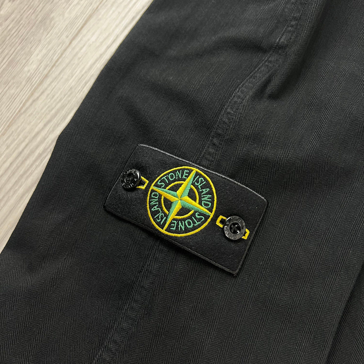 A close up view of the compass badge on the Stone Island Cotton Herringbone Chino Trousers in Black.