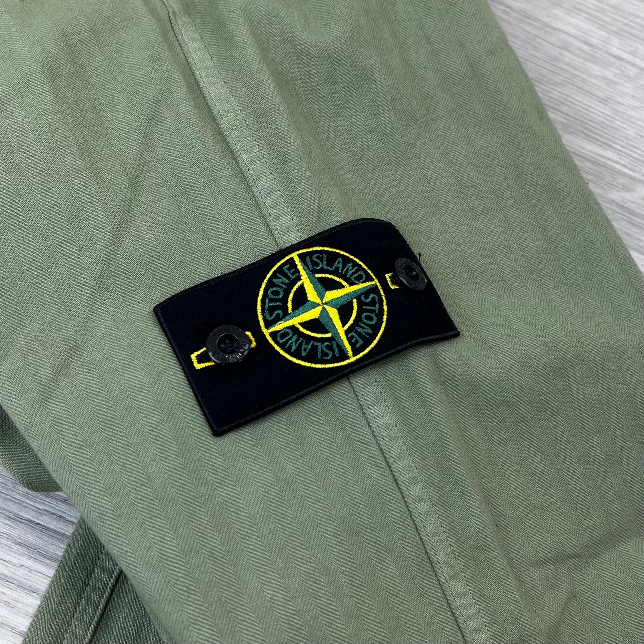 A close up view of the compass badge on the Stone Island Cotton Herringbone Chino Trousers in Laurel Green.